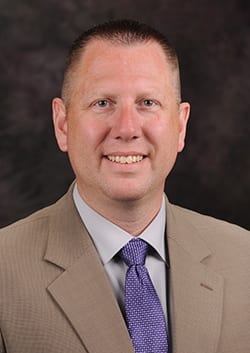 Ulmer posing for headshot in a tan suit jacket and white button up with a purple tie. 