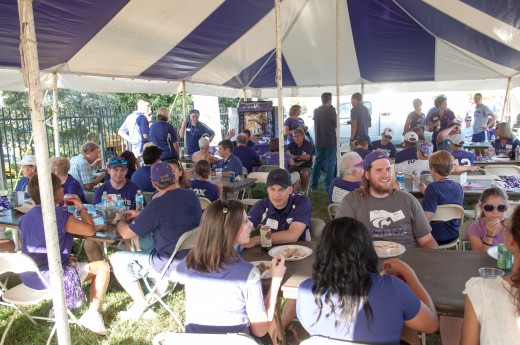 People in purple K-State shirts eating barbecue under a white and purple striped tent.