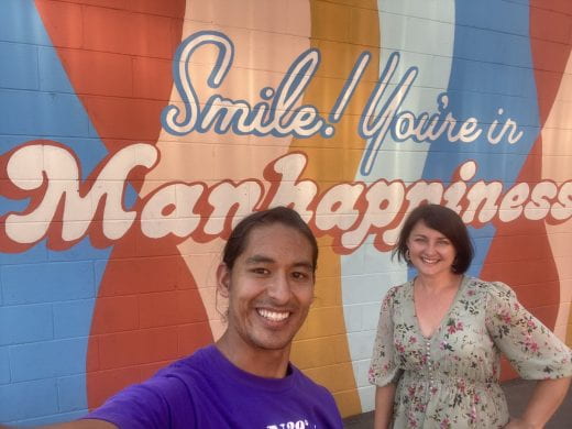 Two K-State students in front of a mural featuring the words "Smile! You're in Manhappiness," in Aggieville, Manhattan, Kansas