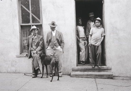 Black and white photograph entitled "Pool Hall (Fort Scott, Kansas)," by Gordon Parks from the collection of the Beach Museum of Art. Showing a group of African American men standing at the door and by the window of a building. Two older men with a dog chatting by the window and three men standing in the doorway.