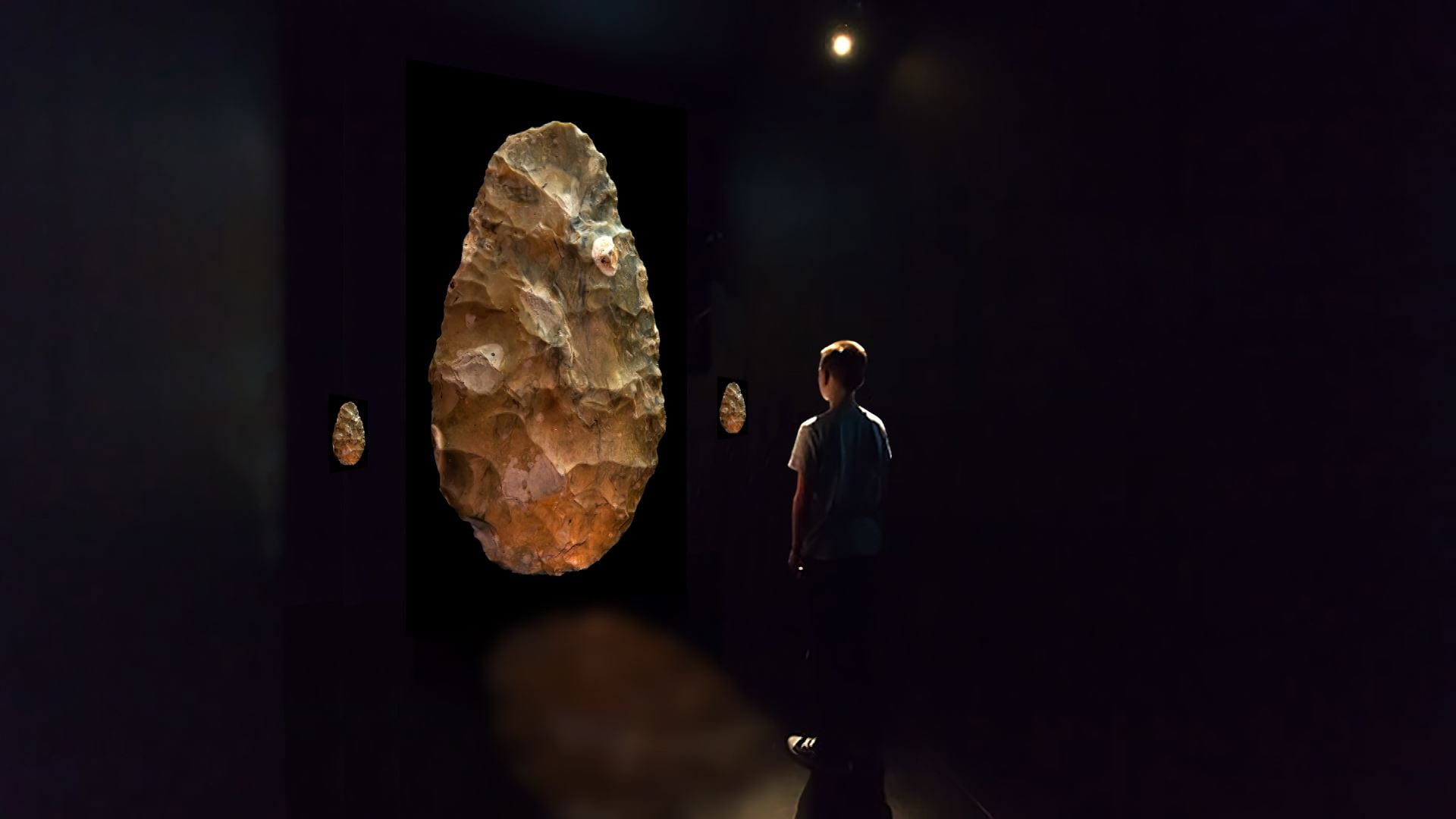 Publicity image for the exhibition "Paleolithic Points from The Forms: Four Worlds | David Lebrun" showing a young boy standing in front of a larger-than-life projected image of an ancient artifact made of stone.